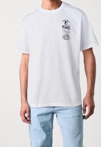 Homme portant un T-shirt blanc avec un logo de diner en cartoon noir et une illustration de pâtes sur la poitrine gauche, associé à un jean bleu clair.