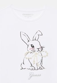 Λευκό T-shirt με μαύρο σχεδιασμό γραμμής ενός κουνελιού διακοσμημένο με σχέδιο που μοιάζει με μαργαριτάρι. Η λέξη "Guess" είναι τυπωμένη σε ασημί κάτω από την εικόνα.
