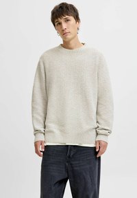 Un pull en tricot beige clair avec un motif texturé, un col et des poignets côtelés, porté par-dessus une chemise blanche, associé à un jean noir décontracté.