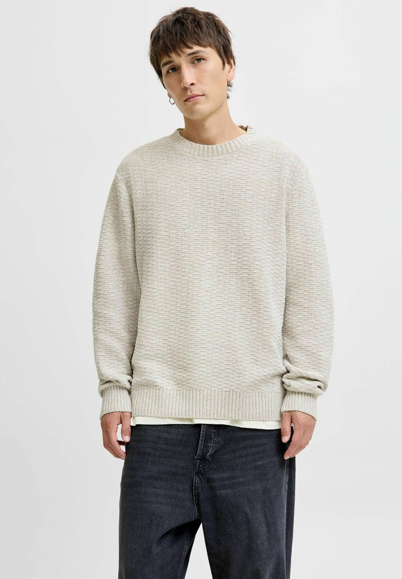 Un pull en tricot beige clair avec un motif texturé, un col et des poignets côtelés, porté par-dessus une chemise blanche, associé à un jean noir décontracté.