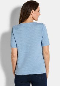 Vrouw met haar rug naar de kijker gekeerd, in een lichtblauwe, textuurshorts met kanten rand en donkere jeans, voor een effen achtergrond.