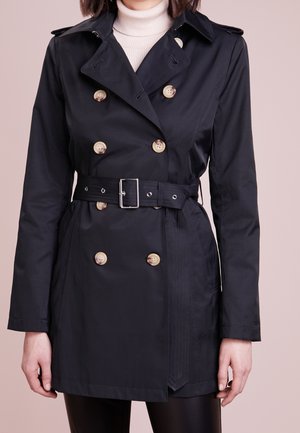 Nainen, jolla on musta kaksirivinen trenssitakki, jossa on beiget napit ja siihen sopiva vyö, valkoisen pooloneuleen päällä.