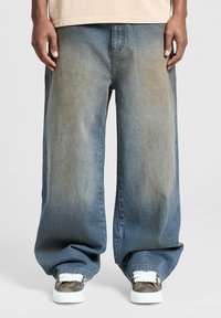 Brede benbukser i blå denim med falmet farve, der har en løs pasform og opkrøllede manchetter. Båret med mønstrede sneakers og en let top.
