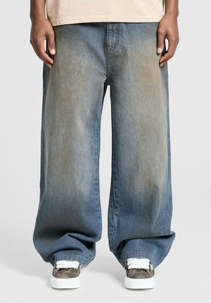 Pantaloni di jeans blu a gamba larga con colorazione sbiadita, caratterizzati da una vestibilità ampia e orli rifiniti. Indossati con sneakers a motivi e una maglietta leggera.