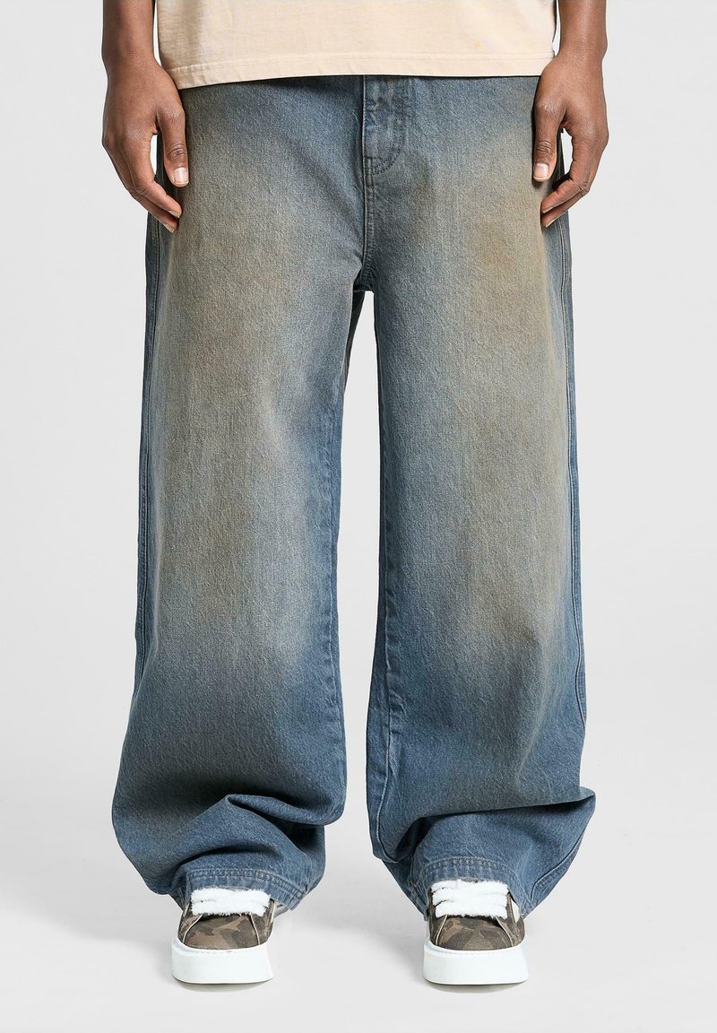 Brede benbukser i blå denim med falmet farve, der har en løs pasform og opkrøllede manchetter. Båret med mønstrede sneakers og en let top.