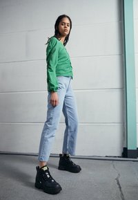 Haut à manches longues vert en tissu texturé, jean droit bleu clair et baskets noires chunky avec des accents jaunes.