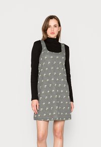 Vestido quadriculado em preto e branco com padrões florais, decote quadrado e alças largas, combinado com uma blusa preta de mangas compridas.