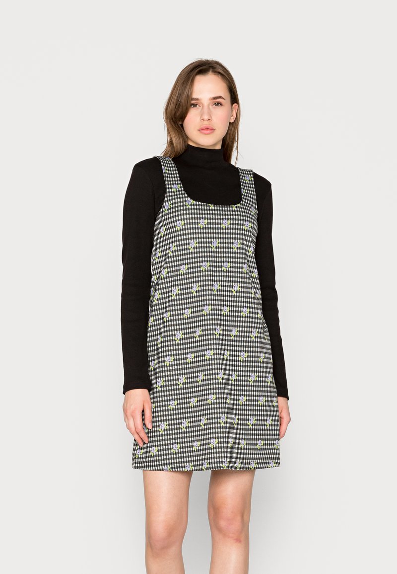 Vestido quadriculado em preto e branco com padrões florais, decote quadrado e alças largas, combinado com uma blusa preta de mangas compridas.