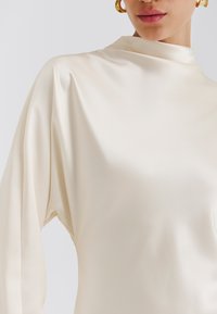 Top de satén color crema con cuello alto y mangas largas y voluminosas, con una textura suave y detalles mínimos.
