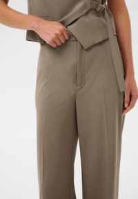 Pantaloni bej, ajustați, cu un front plat, pliu subtil și o buzunar lateral; materialul pare neted, cu un ușor luciu.