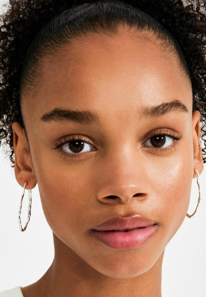 Orecchini a cerchio in argento, design intrecciato, di media dimensione; texture liscia. Ritratto ravvicinato che mette in risalto i tratti del viso.