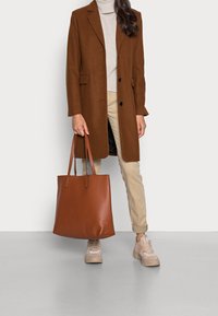 Cappotto di lana marrone, maglione di mohair beige, pantaloni beige e sneakers marroni chiari. Grande borsa tote in pelle marrone con due manici. Texture liscia.