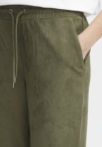 Olivegrønne joggebukser laget av mykt, semsket materiale med elastisk midjebånd, snøring og sidelommer.
