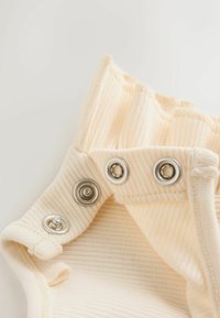 Ljusbeige ribbad body med volangkrage; har silverknappar längst ner för stängning; mjuk textur, slätt tyg.