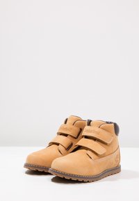 Botas de cuero marrón claro con doble correa de Velcro, puntera redondeada y suela de goma con un patrón de tracción. Marca Timberland en el lateral.