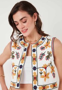 Embroidered sleeveless vest in white with floral patterns in yellow, red, blue, and green. Features a round neckline and open front design.
