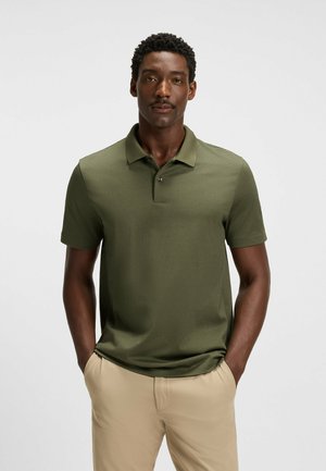 Donkergroene polo met een gestructureerd patroon, korte mouwen en een knoopsluiting met twee knopen. Gecombineerd met lichtbeige broeken.