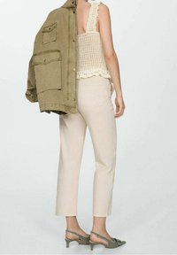 Beige cropped broeken, een crèmekleurig kant tanktop met geschulpte randen, en een olijfgroene oversized jas over de schouder gedrapeerd. Groene hoge hakken sandalen.