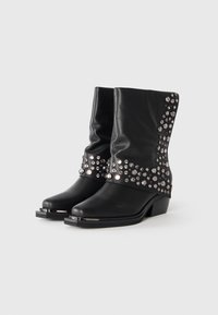 Botas negras de media pantorrilla con tacones apilados, punteras puntiagudas, adornos metálicos alrededor del eje superior y detalles en metal en las capuchas de los dedos.