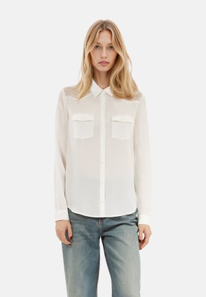 Camicia bianca a maniche lunghe con colletto, due taschini sul petto e dettagli in pizzo trasparente sulle spalle. Tessuto morbido e leggero.