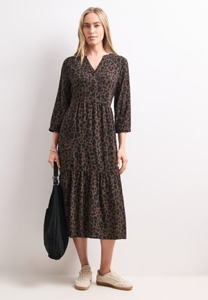 Femme portant une robe midi imprimé léopard marron et noir avec des manches trois-quarts, tenant un sac noir et portant des baskets beiges.