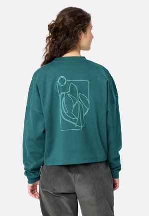 Donna con capelli ricci che indossa una felpa verde acqua con un disegno astratto lineare sulla schiena e pantaloni grigio scuro, in piedi con le spalle rivolte verso di noi.