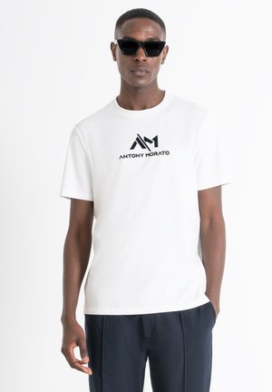 Camiseta blanca de algodón con un logo negro "ANTONY MORATO". El modelo lleva gafas de sol angulares negras, combinadas con pantalones oscuros.
