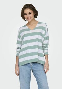 Pull en V rayé vert et blanc avec des manches longues, fabriqué dans un matériau doux. Coupe décontractée avec un ourlet légèrement court.