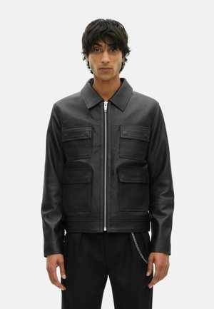 Jeune homme aux cheveux bruns portant une veste en cuir noir avec fermeture éclair à l'avant et quatre poches à rabat, se tenant devant un fond uni.