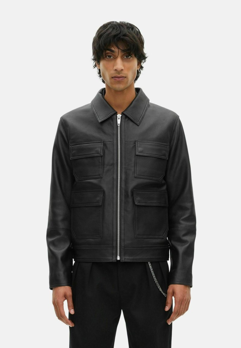 Young man with dark hair wearing a black leather jacket with front zipper and four flap pockets, standing against a plain background.