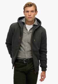 Donkergrijze zip-up hoodie met een gevoerde capuchon, gecombineerd met een geruit overhemd, olijfgroene broek en een zwarte riem. Casual en comfortabele look.