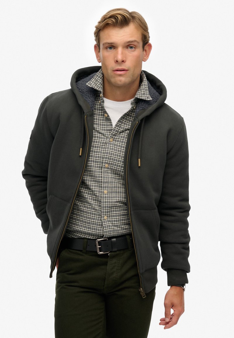 Donkergrijze zip-up hoodie met een gevoerde capuchon, gecombineerd met een geruit overhemd, olijfgroene broek en een zwarte riem. Casual en comfortabele look.