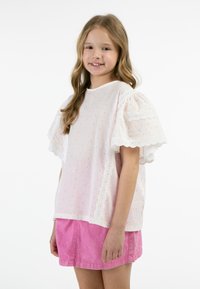 Blusa bianca con pois rosa e maniche a sbuffo con bordino in pizzo, abbinata a una gonna rosa. Il tessuto appare leggero e dalla texture interessante.