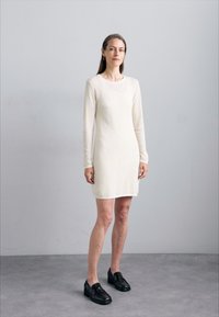 Abito in maglia a maniche lunghe color crema con scollo rotondo, taglio aderente e texture liscia, abbinato a mocassini neri.