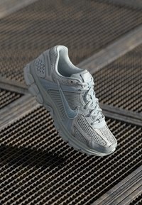 Ljust grå Nike löparskor med nätpaneler och snören som vilar på en texturerad metallyta.