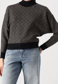 Pull noir tricoté avec motif géométrique doré et col montant. Poignets et ourlet côtelés, coupe décontractée, porté avec un denim bleu clair.