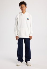 White hoodie with front pocket, featuring a colorful graphic; paired with navy blue pants and white sneakers. Smooth texture, casual design.