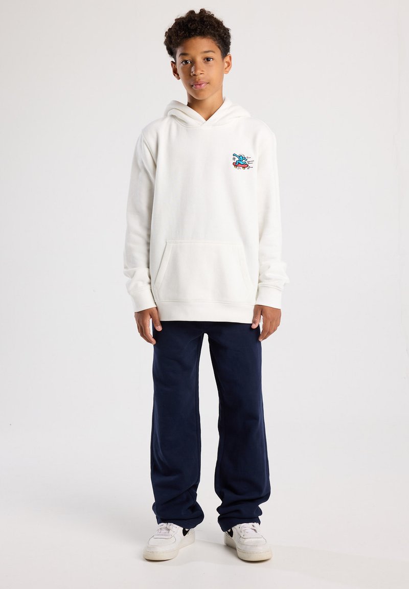 White hoodie with front pocket, featuring a colorful graphic; paired with navy blue pants and white sneakers. Smooth texture, casual design.