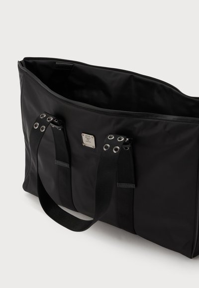 Sac cabas rectangulaire noir avec double poignée comportant des œillets métalliques et une petite plaque de marque métallique carrée sur le devant, ouverture supérieure avec fermeture éclair.