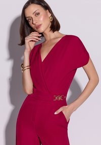 Fuksjowy kombinezon z głębokim dekoltem w kształcie litery V, krótkimi rękawami i ozdobną złotą klamrą w talii. Gładka tkanina o eleganckim fasonie.