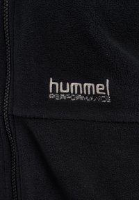 Svart fleecejacka med hela dragkedjan framtill, mjuk textur och en vit broderad logotyp med texten "hummel PERFORMANCE."