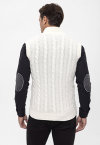 Weißer Kabelstrick-Weste mit hohem Kragen, mit einem strukturierten Muster. Getragen über einem schwarzen Langarmshirt mit Ellenbogenpatches.