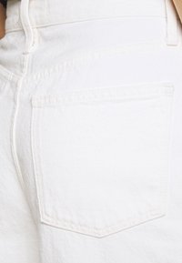 Bílé džíny mají jednu zadní kapsu s tónovým prošitím a jemnou texturou, zobrazené z blízkého bočního pohledu.