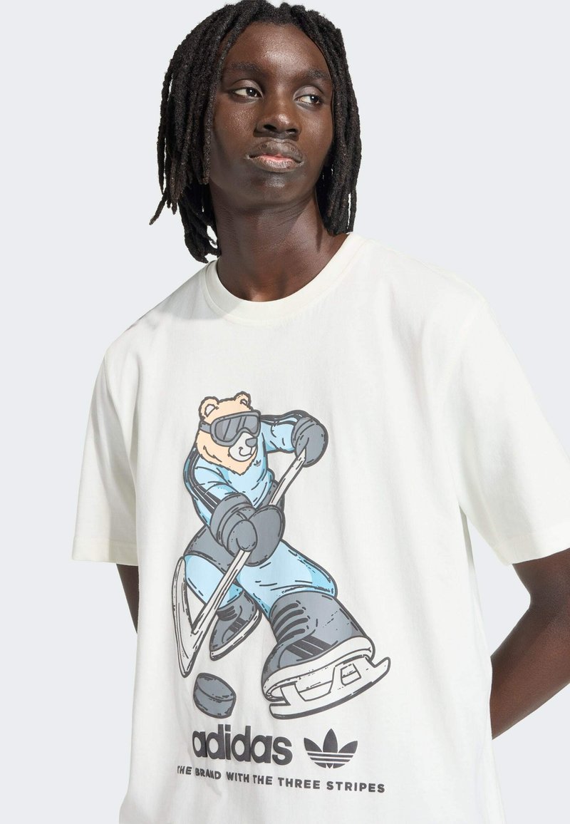 Krämvit bomulls-T-shirt med en tecknad björn i skidkläder som håller en hockeyklubba. Svart broderad logotyp längst ner.