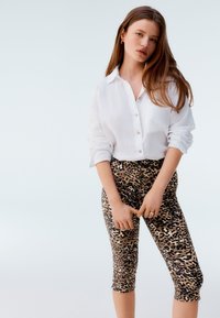 Biała lniana koszula zapinana na guziki w połączeniu z dopasowanymi legginsami o długości do kolan w panterkę, gładkie w fakturze, z brązowo-czarnym wzorem.