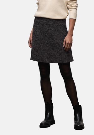 Person trägt einen beigefarbenen Pullover, schwarzen strukturierten A-Linien-Rock, durchsichtige schwarze Strumpfhosen und glänzende schwarze Stiefeletten vor einem schlichten Hintergrund.