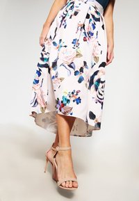 Blommig midikjol i ljusrosa tyg med blå och lila blommor, hög-låg fåll, matchad med nudefärgade sandaletter med remmar.