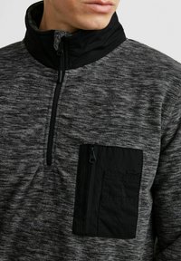 Grijze en zwarte half-zip pullover met een gestructureerd lichaam, hoge kraag en een ritszak op de linkerborst.