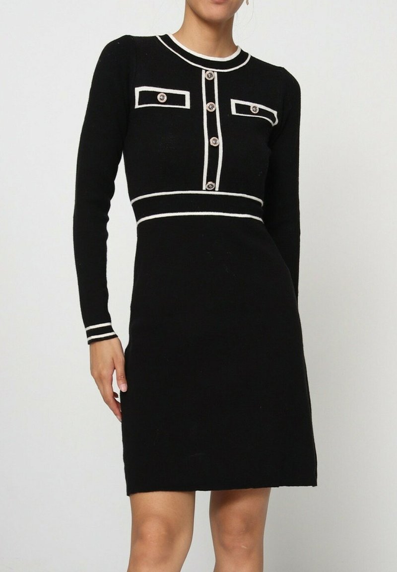 Robe en tricot noire à manches longues, avec des accents blancs, deux poches poitrine et une fermeture à boutons sur le devant. Texture lisse, silhouette ajustée.