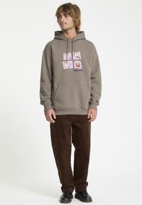 Sudadera gris con estampado gráfico que presenta cuatro caras de dibujos animados. Combinada con pantalones de pana marrón y zapatos negros. Textura suave, corte relajado.